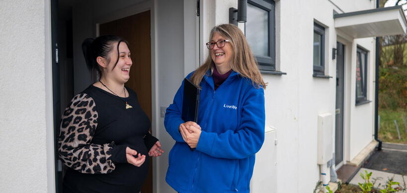 A woman wearing a blue LiveWest jacket is standing outside a house, smiling and talking with another woman who is standing in the doorway.