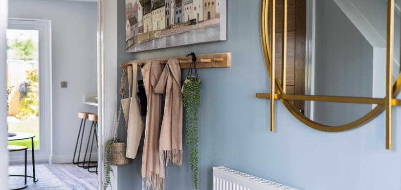 The image shows a tidy hallway with coats and plants hanging on hooks by a wall decorated with a round mirror and a painting of buildings by the water.