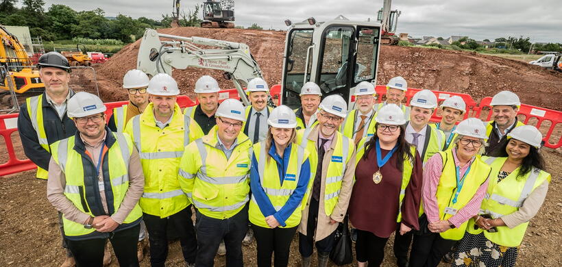 A group of people in hard hats and high-visibility vests pose together on a muddy construction site with excavators.