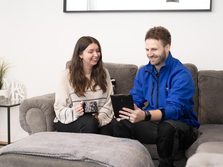 A man and a woman are sitting on a sofa in a living room, with the man holding a tablet and the woman holding a mug as they talk.