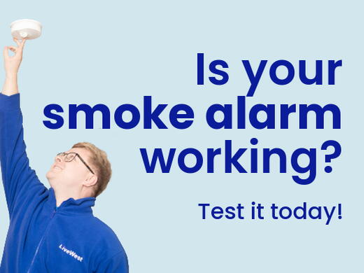 A person wearing glasses and a blue LiveWest jacket is pressing a smoke alarm on the ceiling in a kitchen.
