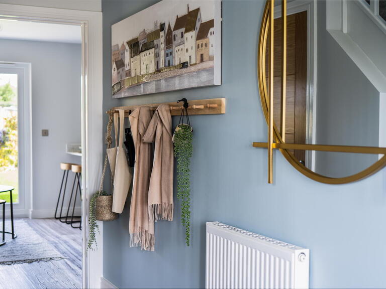 The image shows a tidy hallway with coats and plants hanging on hooks by a wall decorated with a round mirror and a painting of buildings by the water.