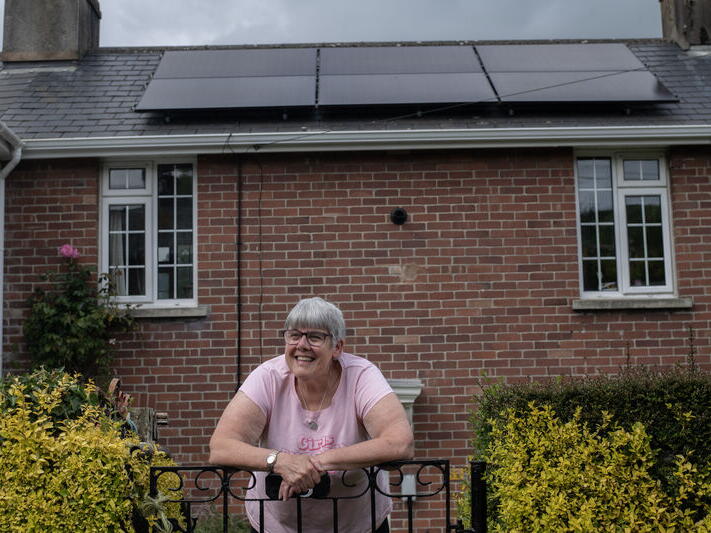 A smiling woman leaning on a garden gate in front of a house with solar panels.