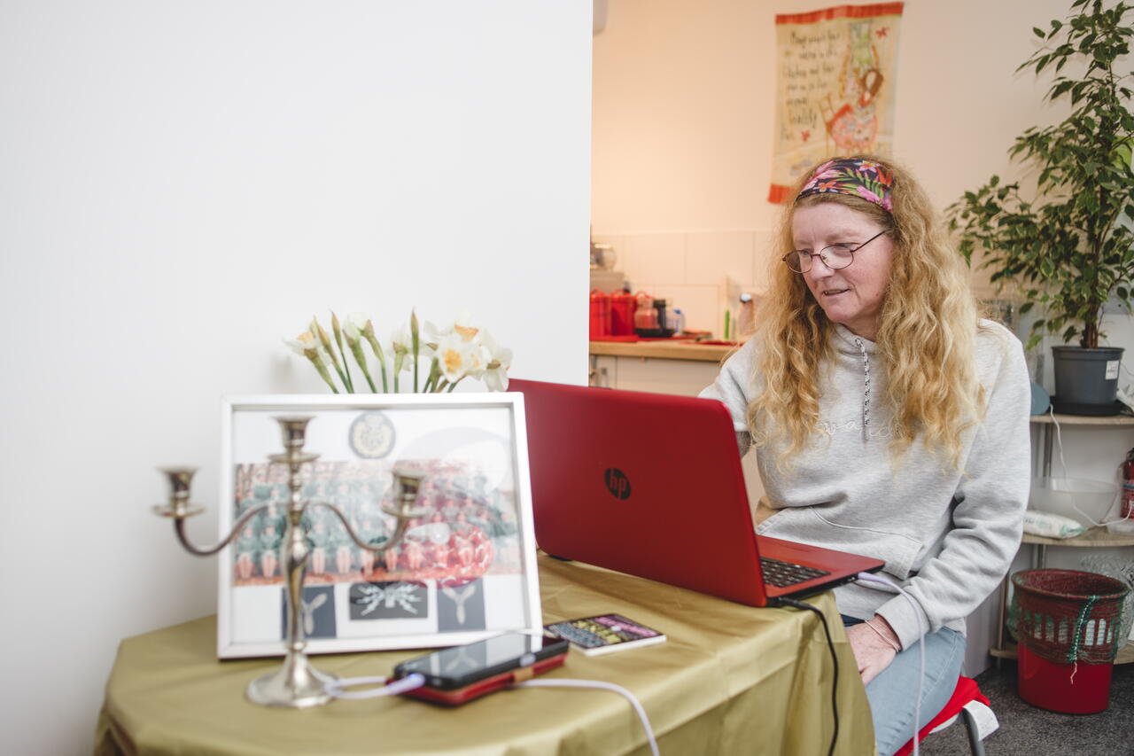 A woman with long hair and glasses is sitting on a red chair, using a red laptop at a table covered with a gold cloth in a cosy indoor setting.