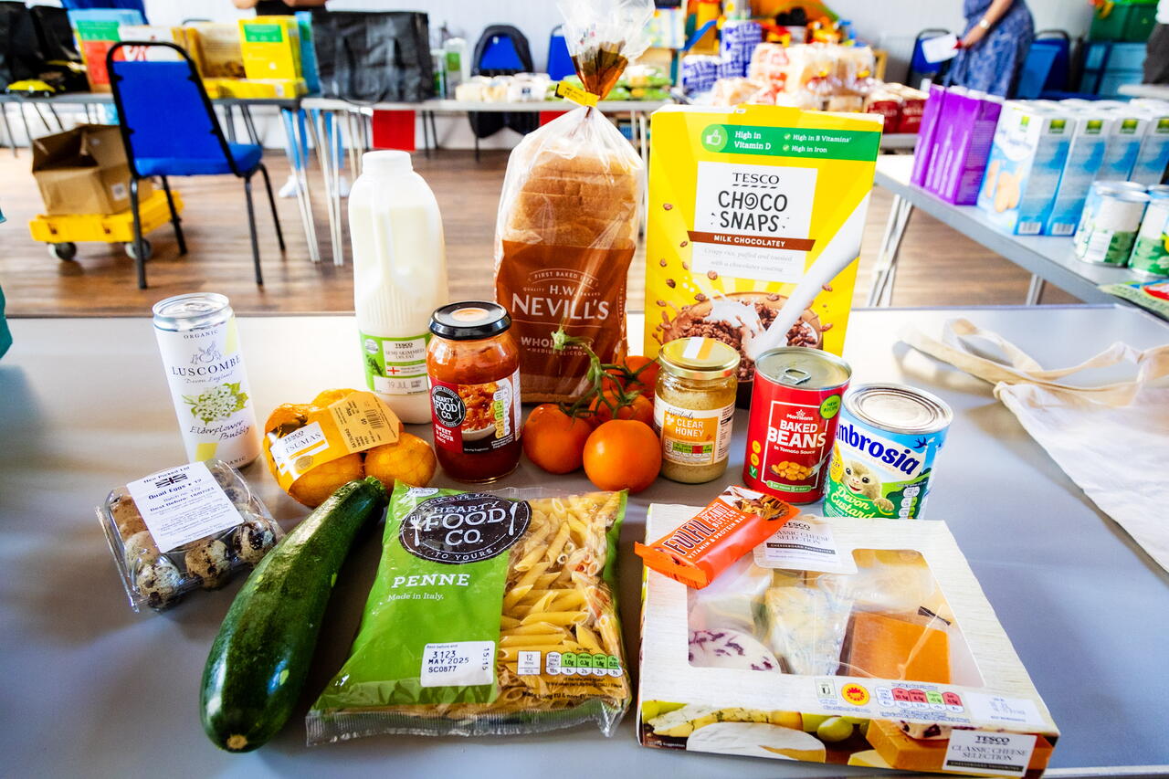 The main subject of the image is a variety of groceries arranged on a table inside a communal hall, suggesting a food distribution or collection setting.