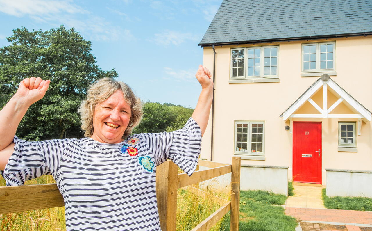A person is smiling and raising their arms in celebration outside a house with a red door on a clear day.