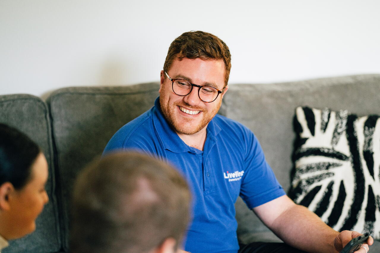 A man wearing glasses and a blue polo shirt smiles while talking to two other people in a living room.