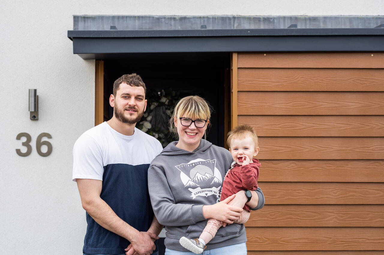 A couple is standing outside the door of their home, with one person holding a baby.
