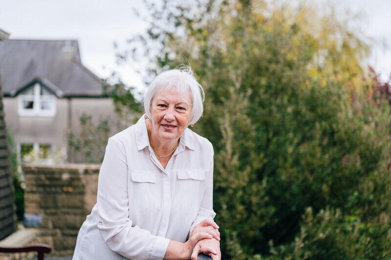 A smiling person with white hair leans on a railing outside in a garden with houses and trees in the background.
