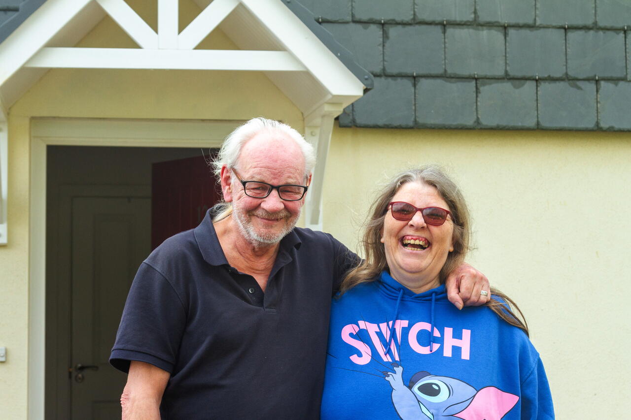 Image shows a couple smiling in front of a house.