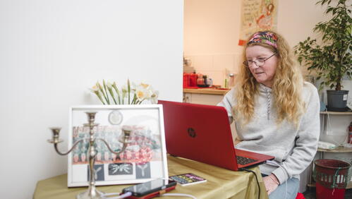 A woman with long hair and glasses is sitting on a red chair, using a red laptop at a table covered with a gold cloth in a cosy indoor setting.