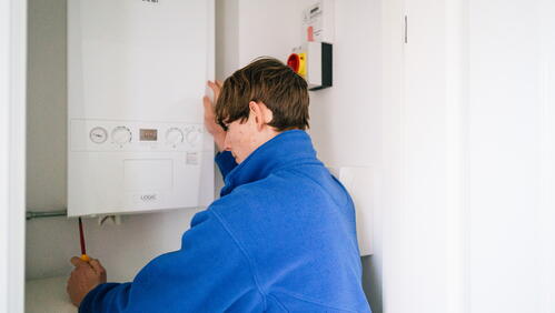 A person is fixing or checking a wall-mounted boiler in a small, white room.