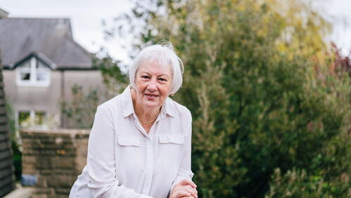 A smiling person with white hair leans on a railing outside in a garden with houses and trees in the background.