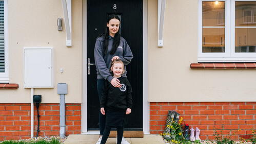 A woman stands behind a child outside the front door of a house, both smiling at the camera.